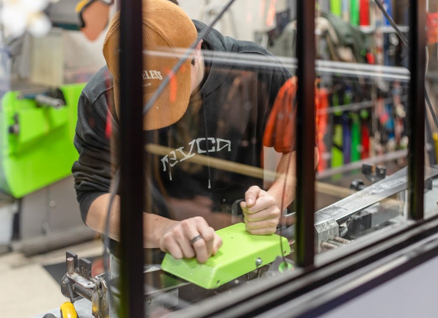 Un homme cire la base d'un ski avec un bloc vert fluorescent dans un atelier de réparation.
