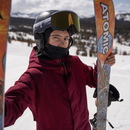 Jeune skieur avec casque et masque, tenant des skis Atomic sur une montagne enneigée.