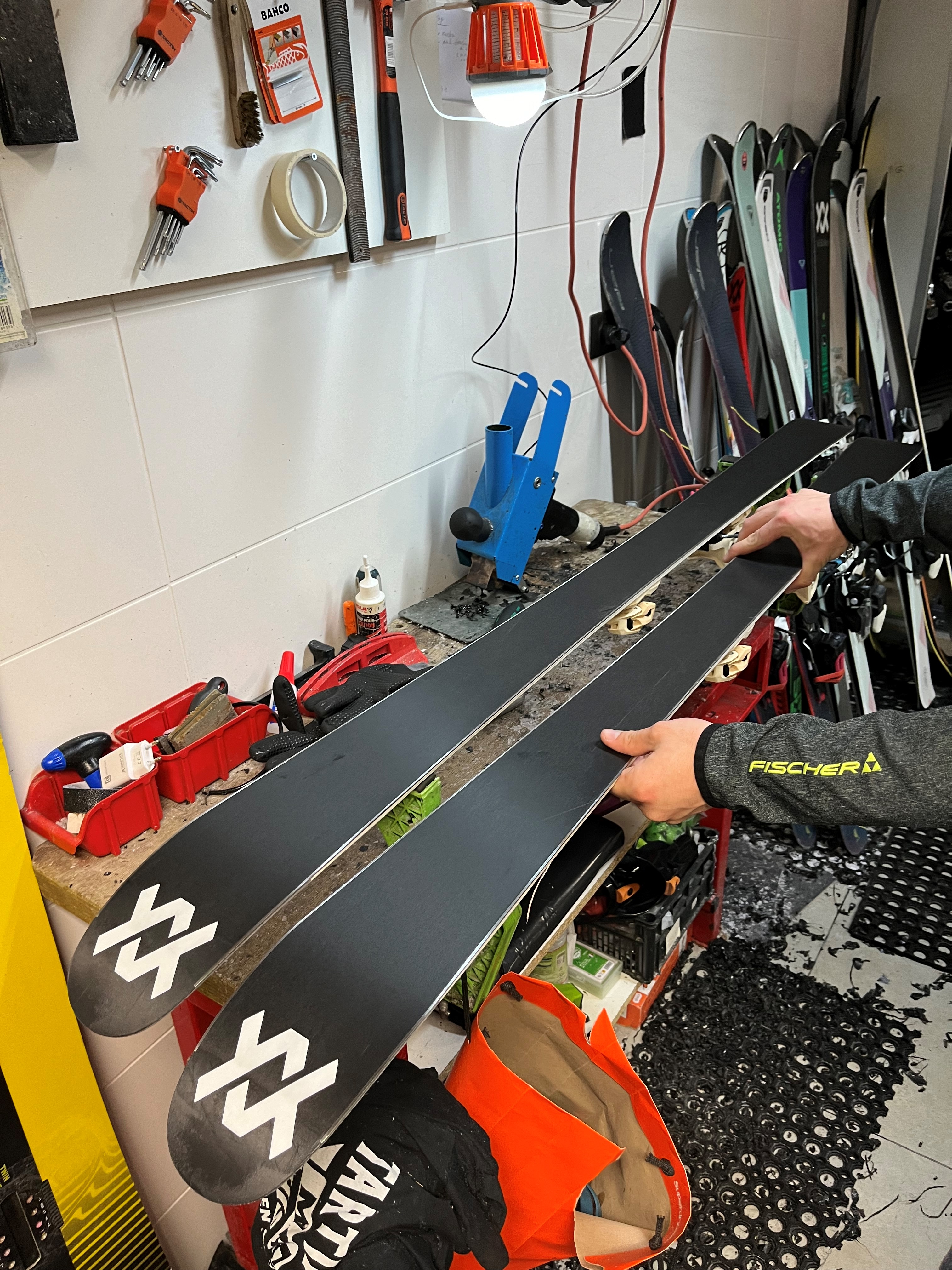Mains tenant une paire de skis noirs Völkl fraîchement fartés, dans un atelier de réparation avec des outils et d'autres skis.