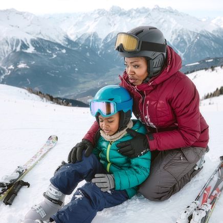 Une femme en tenue de ski réconforte un enfant assis dans la neige. Skis et montagnes enneigées en arrière-plan.