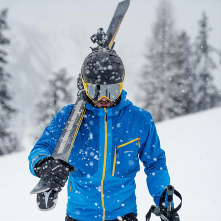 Skieur en veste bleue, casque et masque, portant ses skis sur l'épaule sous la neige tombante en montagne.