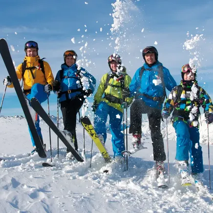 Cinq skieurs joyeux dans la neige fraîche avec de la poudreuse projetée en l'air, sur un sommet montagneux sous un ciel bleu.