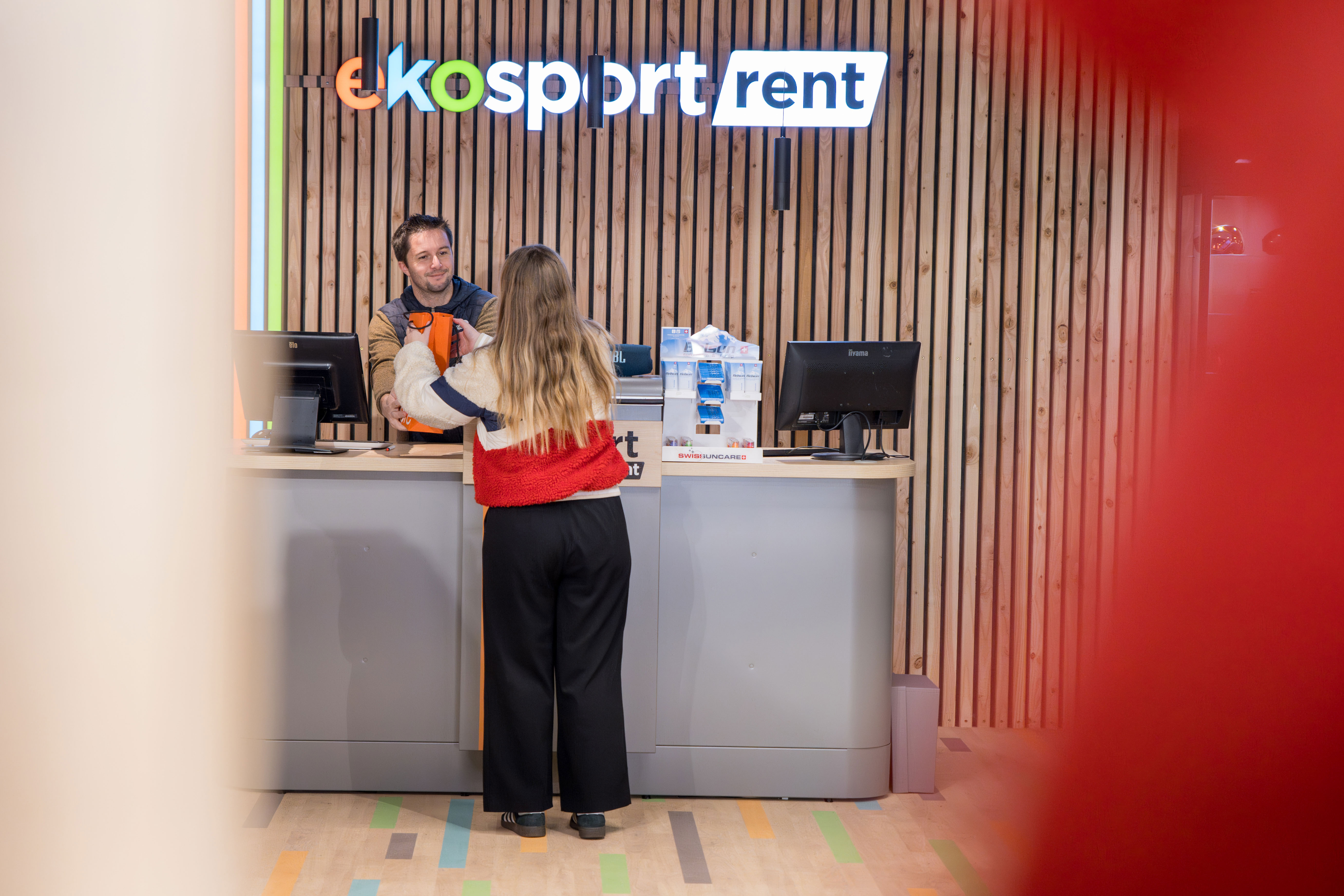 Une femme vue de dos reçoit un article orange d'un employé souriant au comptoir d'Ekosport Rent, devant un mur en bois.