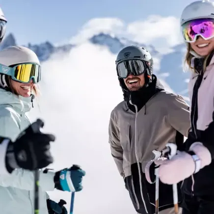 Quatre skieurs souriants avec casques et masques, bavardant sur une montagne enneigée avec des sommets nuageux en arrière-plan.