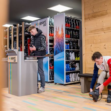 Deux hommes dans un magasin de location de ski. L'un ajuste ses fixations sur un comptoir, l'autre enfile ses chaussures de ski sur un banc.