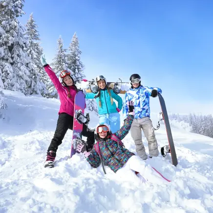 Quatre jeunes snowboarders souriants posent dans un paysage de montagne enneigé, sous un ciel bleu éclatant.
