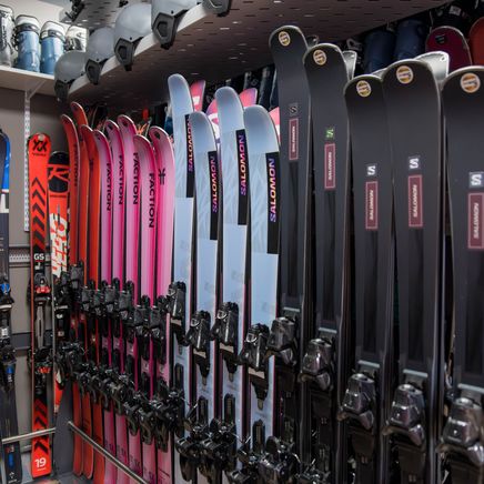 Nombreuses paires de skis de marques et couleurs variées (noir, rose, rouge, bleu) alignées dans un magasin, avec bottes et casques.