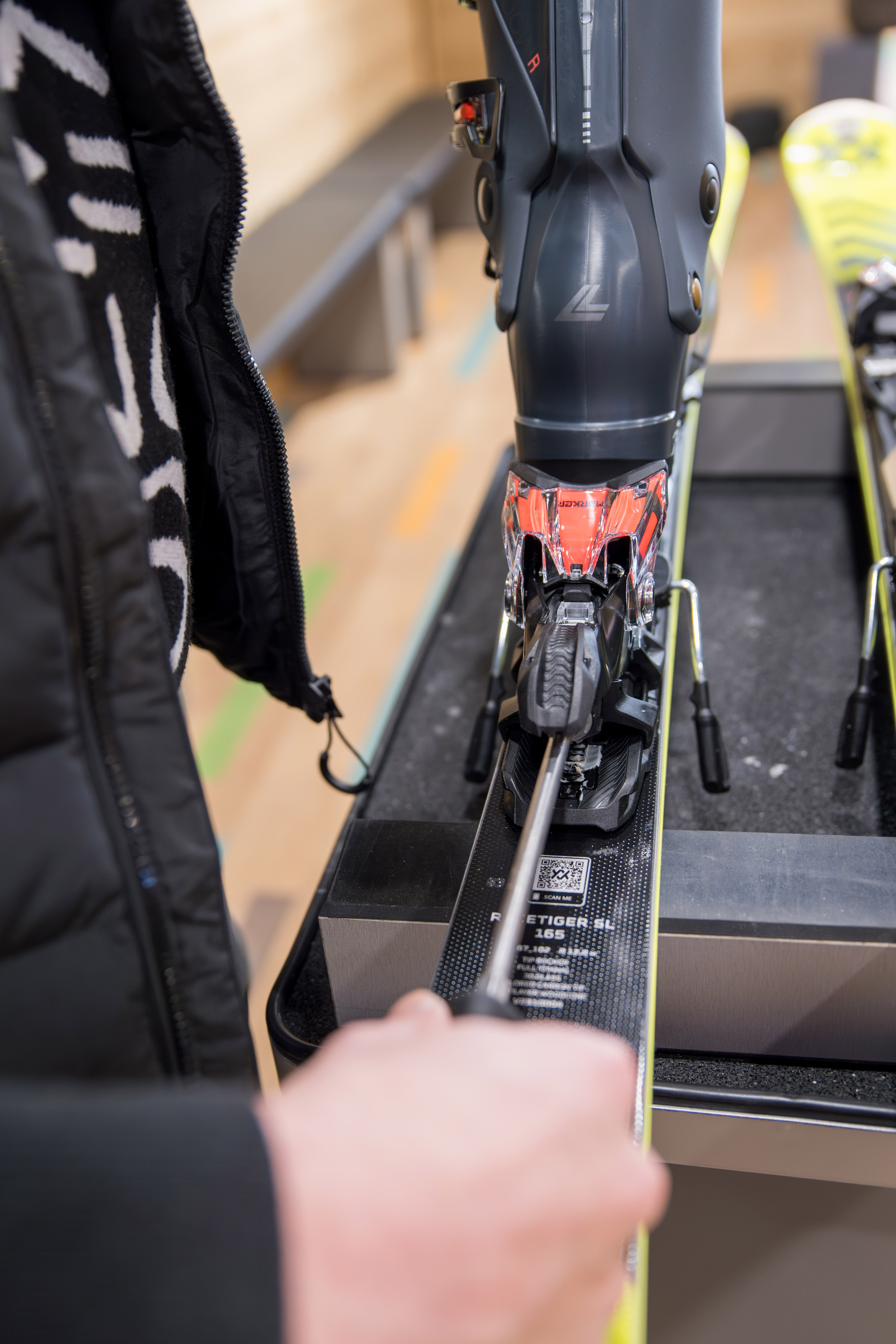 Main ajustant une fixation de ski noire et rouge avec une chaussure de ski gris foncé insérée, sur un banc de réglage.