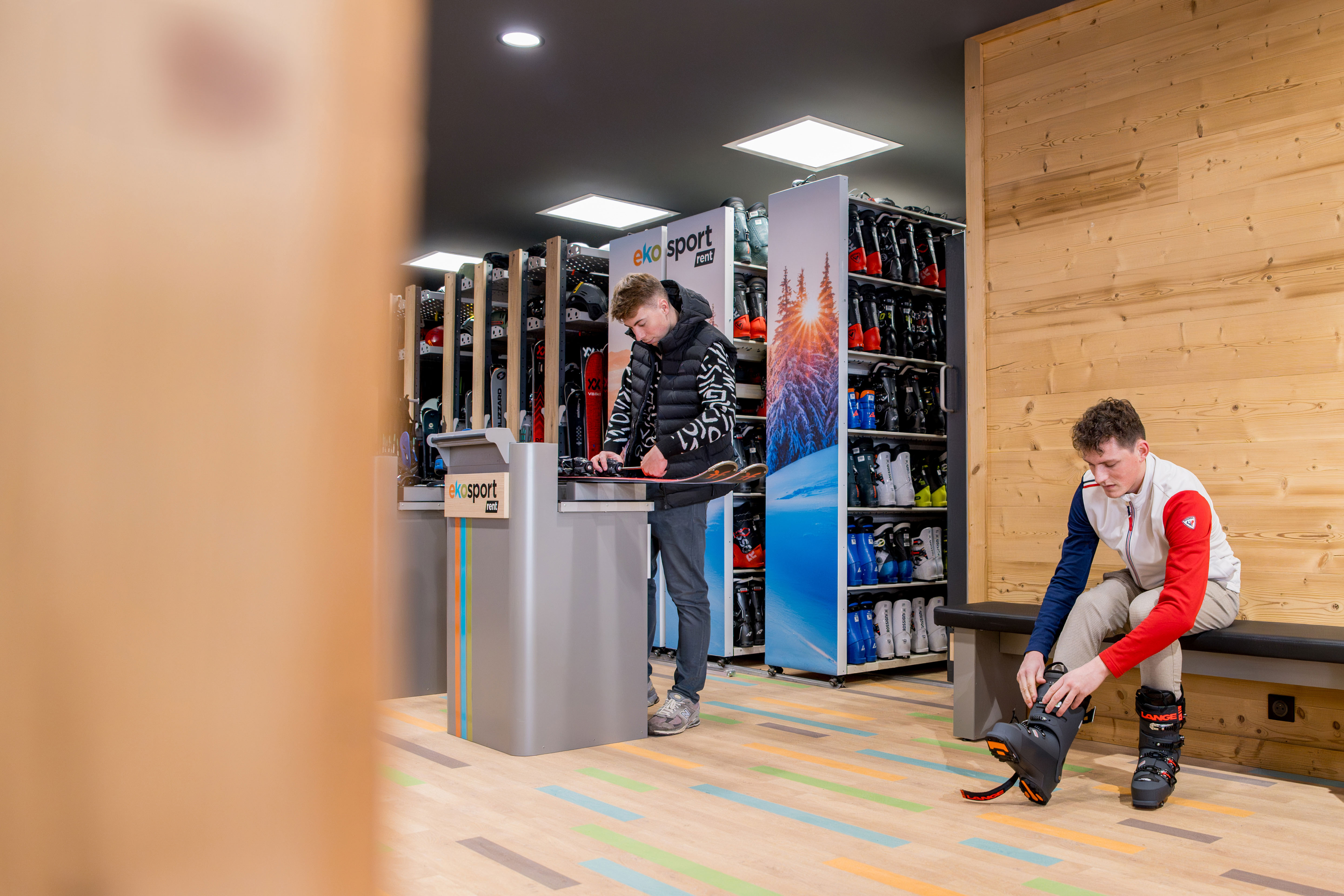 Deux jeunes hommes dans un magasin de location de skis. L'un ajuste ses chaussures de ski, l'autre tient des skis à un comptoir.