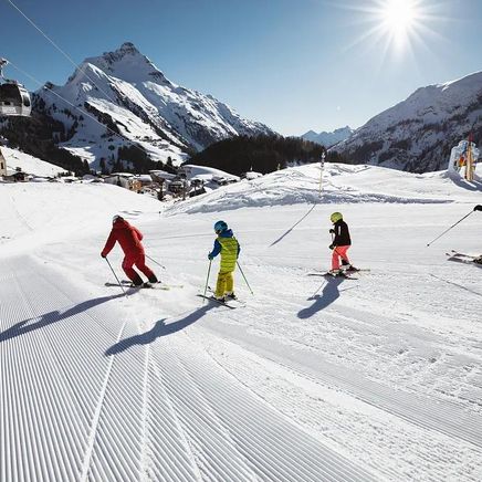 Une famille de quatre skieurs descend une piste damée sous un soleil éclatant, avec des montagnes et un village alpin en fond.