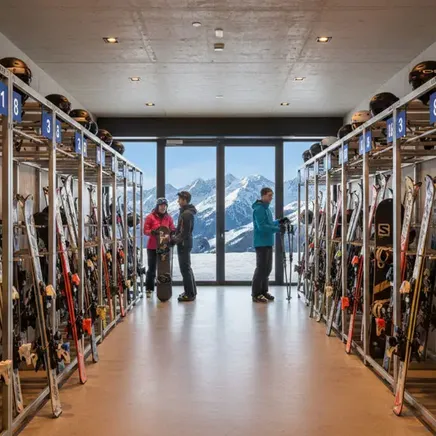 Local à skis et casques avec rangées d'équipement. Trois personnes devant une baie vitrée panoramique sur des montagnes enneigées.